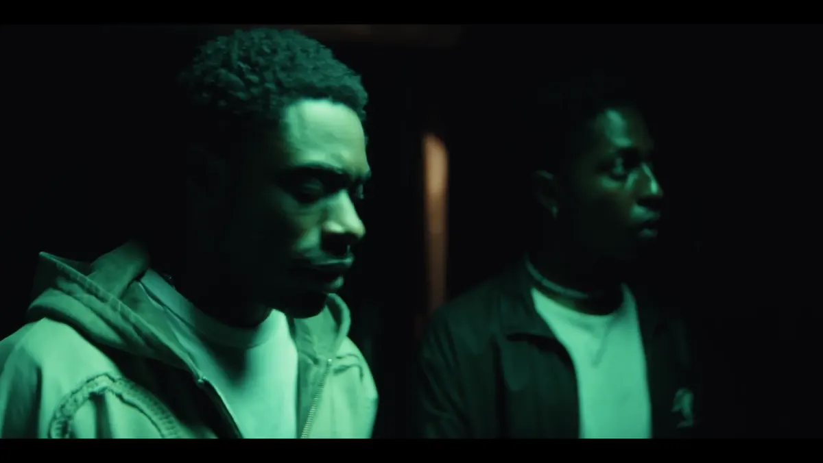 Two men stand together in a dark green-lit interior.